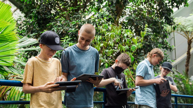 Five young people looking down at tablets, with the trees of the botanic garden in the background