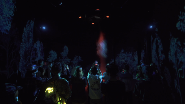 A circle of people viewed from behind, interacting with an image projected onto smoke.