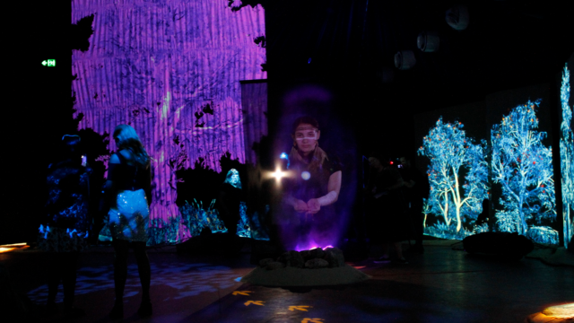A dark indoor scene with a projected purple tree on the left and teal trees on the right. The image of a First Nations member is projected above a purple fire, reaching out to the viewer.
